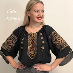 Pu&ograve; includere: Blusa nera ricamata con filo dorato e un fiocco rosso. La blusa ha una scollatura rotonda e maniche a tre quarti.
