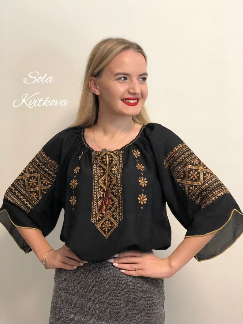 Pu&ograve; includere: Blusa nera ricamata con filo dorato e un fiocco rosso. La blusa ha una scollatura rotonda e maniche a tre quarti.