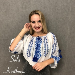 May include: A woman wearing a white blouse with blue embroidery and a grey skirt. The blouse has a tassel detail and the text "Sola Kvitkova" is written below the woman.