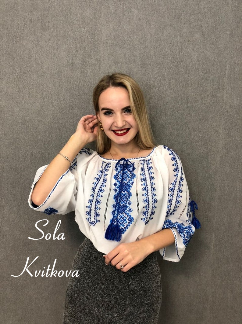 May include: A woman wearing a white blouse with blue embroidery and a grey skirt. The blouse has a tassel detail and the text "Sola Kvitkova" is written below the woman.