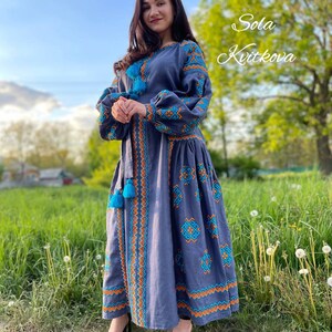 May include: A woman wearing a long, blue linen dress with embroidered details in orange and turquoise. The dress has a fitted bodice and a flowing skirt. The woman is standing in a field with green grass and dandelions. The text "Sola Kvitkova" is written in white on a green background in the upper right corner of the image.