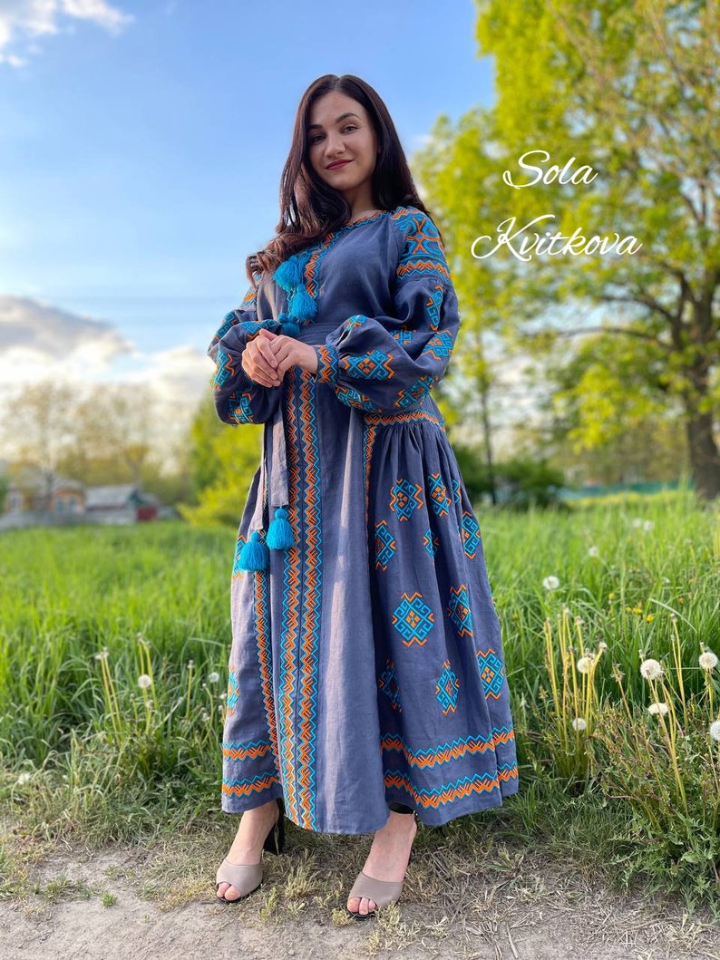 May include: A woman wearing a long, blue linen dress with embroidered details in orange and turquoise. The dress has a fitted bodice and a flowing skirt. The woman is standing in a field with green grass and dandelions. The text "Sola Kvitkova" is written in white on a green background in the upper right corner of the image.