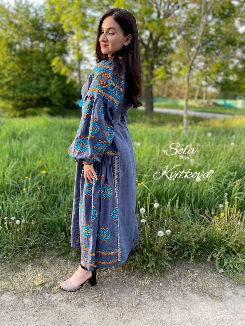 May include: A woman wearing a long, gray linen dress with blue and orange embroidered patterns. The dress has a loose fit and long sleeves. The woman is standing in a grassy field with trees in the background. The text "Sola Kvitkova" is written in cursive on the right side of the image.