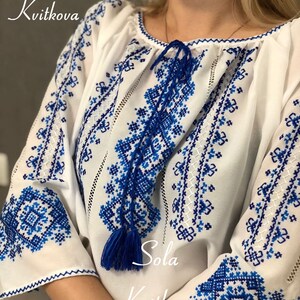 May include: A white embroidered blouse with blue floral patterns. The blouse has a gathered neckline and long sleeves with blue tassels. The text "Sola Kvitkova" is embroidered on the front of the blouse.