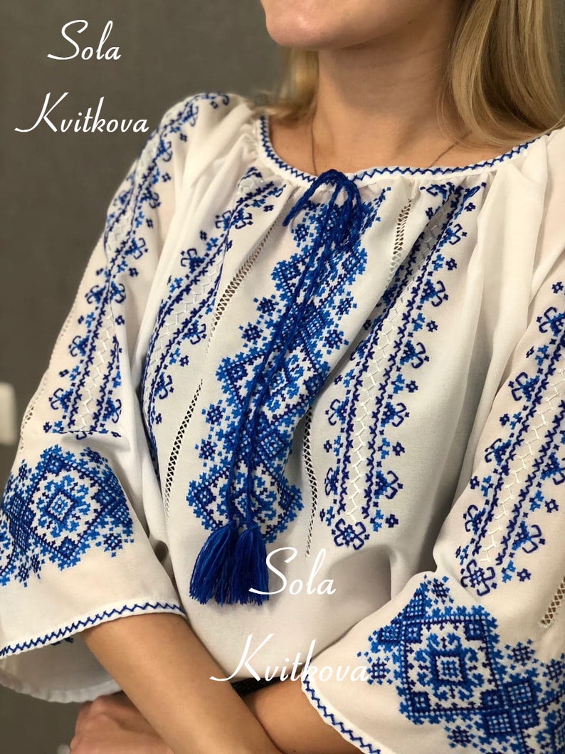 May include: A white embroidered blouse with blue floral patterns. The blouse has a gathered neckline and long sleeves with blue tassels. The text "Sola Kvitkova" is embroidered on the front of the blouse.