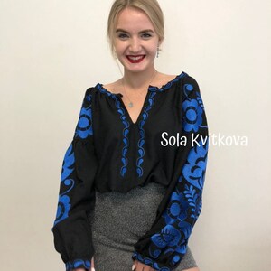 Op de afbeelding: Een zwarte geborduurde blouse met blauwe bloemenmotieven. De blouse heeft een V-hals en lange mouwen. De tekst "Sola Kvitkova" is zichtbaar aan de rechterkant van de blouse.