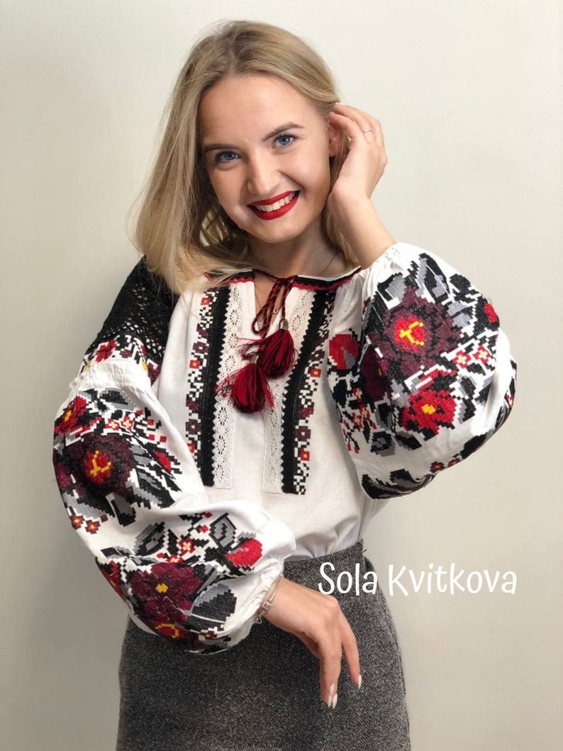 May include: A white embroidered blouse with red, black, and gray floral patterns. The blouse has long sleeves with a gathered cuff and a round neckline. The neckline features a red tassel. The text "Sola Kvitkova" is visible in the bottom right corner of the image.