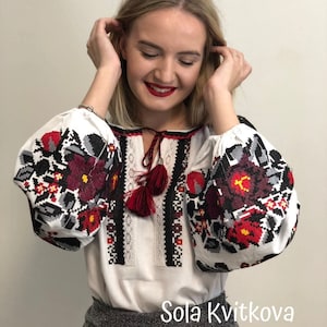 May include: A white embroidered blouse with red and black floral patterns. The blouse has a V-neckline and long sleeves. The sleeves have a gathered look at the shoulders. The blouse has a red tassel tie at the neckline. The text "Sola Kvitkova" is visible at the bottom of the image.