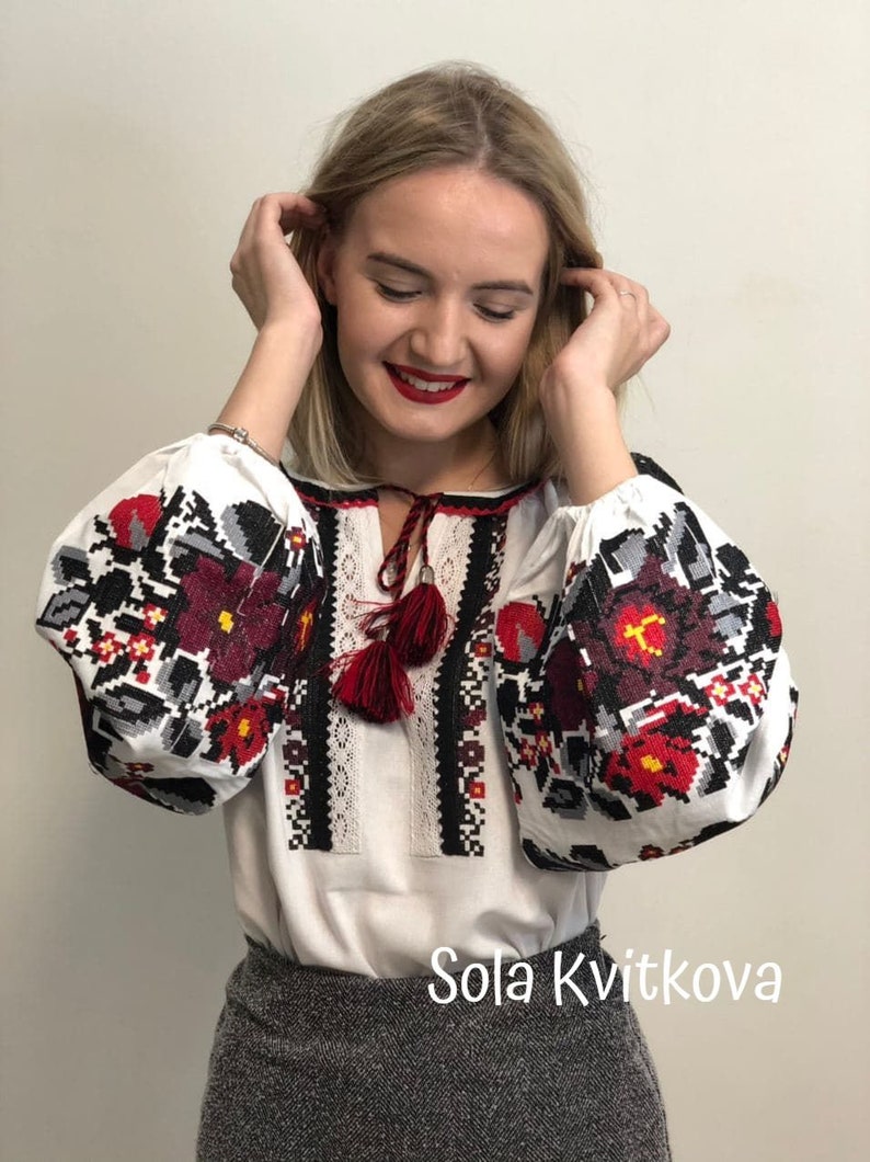 May include: A white embroidered blouse with red and black floral patterns. The blouse has a V-neckline and long sleeves. The sleeves have a gathered look at the shoulders. The blouse has a red tassel tie at the neckline. The text "Sola Kvitkova" is visible at the bottom of the image.