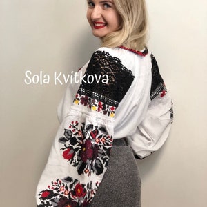 May include: A white embroidered blouse with black lace detailing on the shoulders and sleeves. The blouse features a floral pattern in red, black, and yellow. The text "Sola Kvitkova" is visible on the front of the blouse.