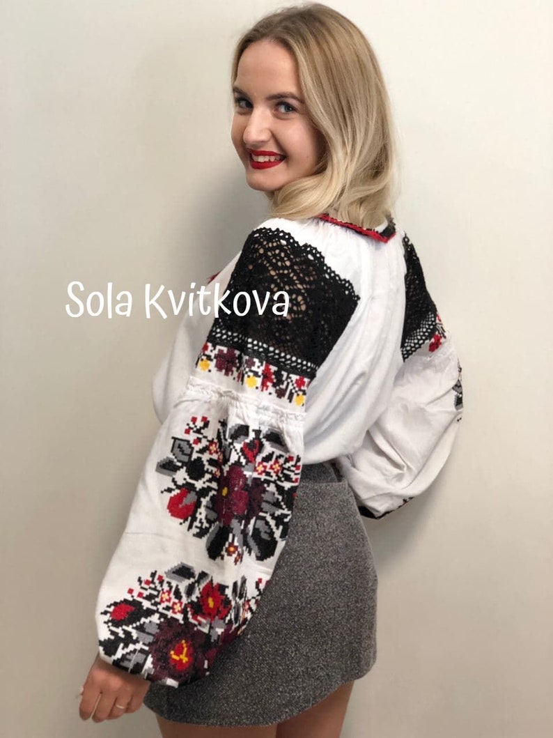 May include: A white embroidered blouse with black lace detailing on the shoulders and sleeves. The blouse features a floral pattern in red, black, and yellow. The text "Sola Kvitkova" is visible on the front of the blouse.