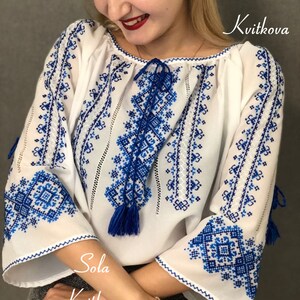 May include: White embroidered blouse with blue geometric patterns. The blouse has a gathered neckline and long sleeves with blue tassels. The text "Sola Kvitkova" is visible on the bottom of the image.