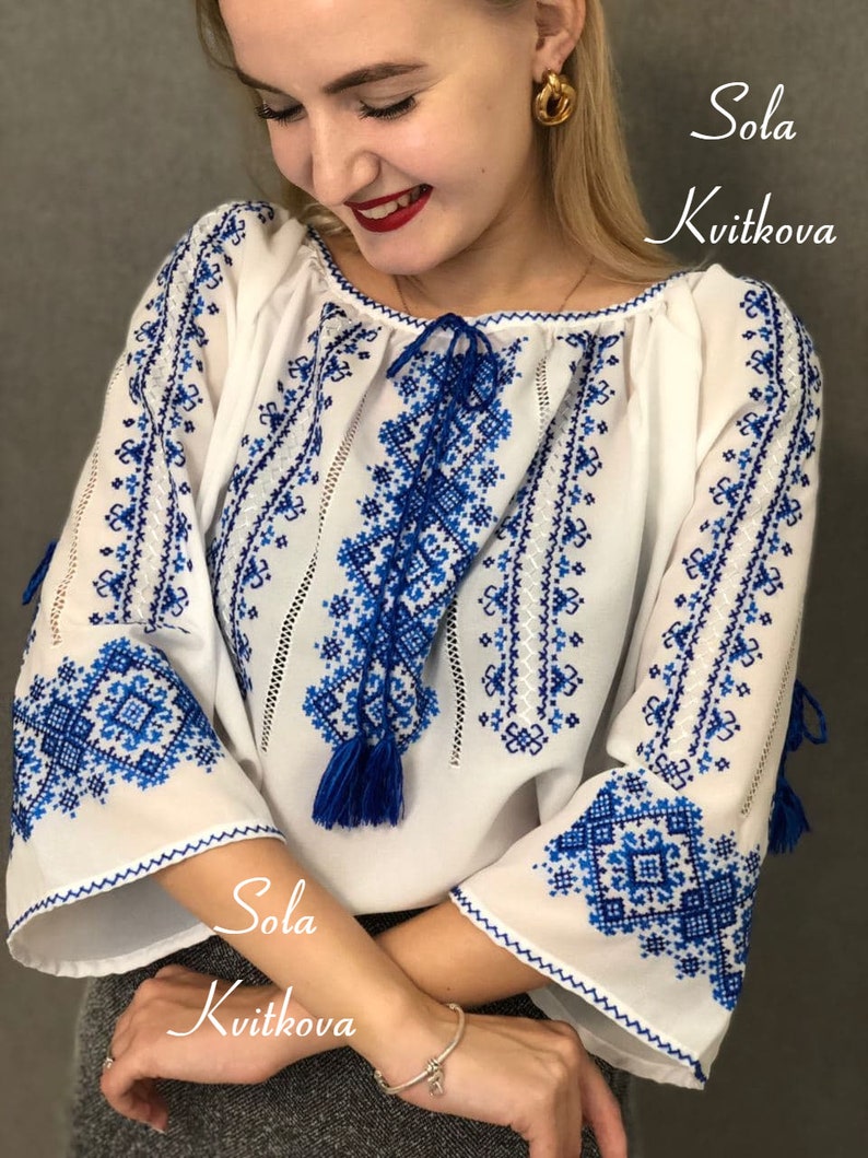 May include: White embroidered blouse with blue geometric patterns. The blouse has a gathered neckline and long sleeves with blue tassels. The text "Sola Kvitkova" is visible on the bottom of the image.