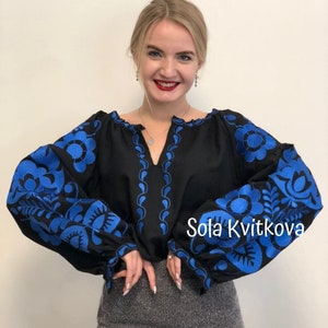 Op de afbeelding: Een zwarte geborduurde blouse met blauwe bloemenpatronen op de mouwen en de halslijn. De tekst "Sola Kvitkova" is zichtbaar in de rechterbenedenhoek van de afbeelding.