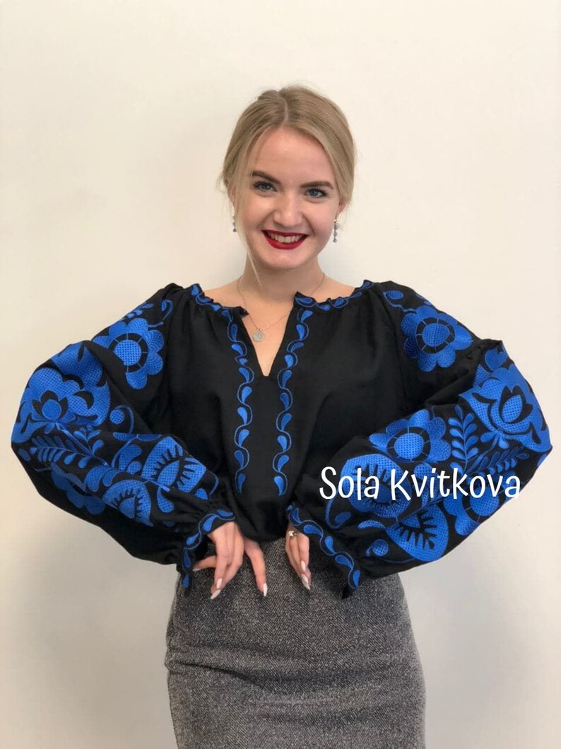 Op de afbeelding: Een zwarte geborduurde blouse met blauwe bloemenpatronen op de mouwen en de halslijn. De tekst "Sola Kvitkova" is zichtbaar in de rechterbenedenhoek van de afbeelding.