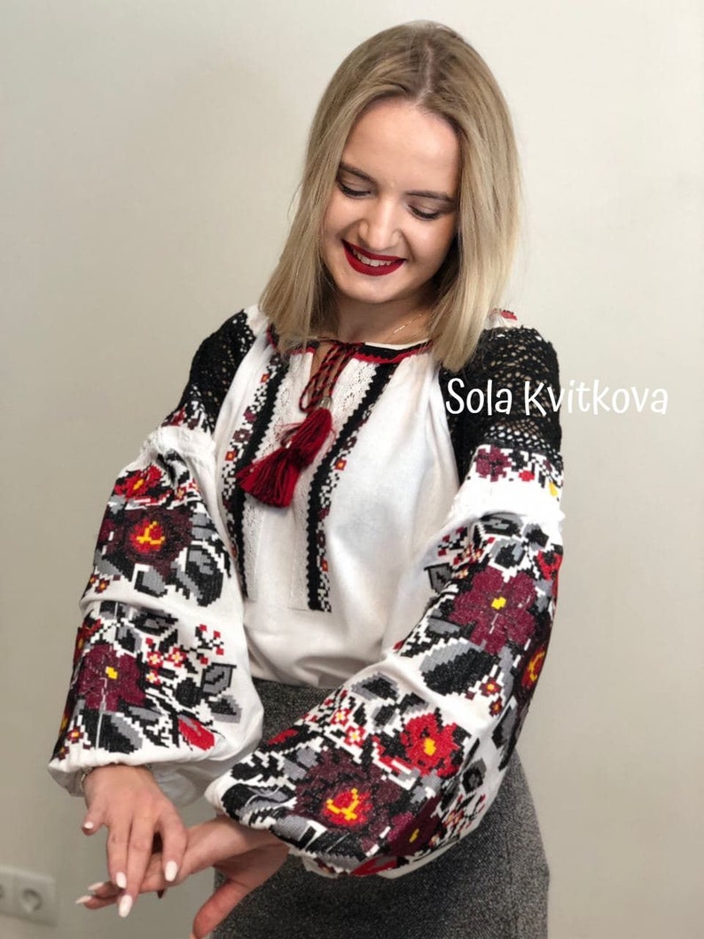 May include: White embroidered blouse with red and black floral patterns. The sleeves have a black lace trim. The blouse has a red tassel hanging from the neckline. The text "Sola Kvitkova" is visible in the upper right corner of the image.