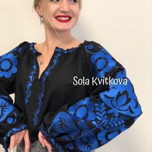 Op de afbeelding: Een zwarte geborduurde blouse met blauwe bloemenmotieven. De tekst "Sola Kvitkova" is zichtbaar aan de rechterkant van de blouse.
