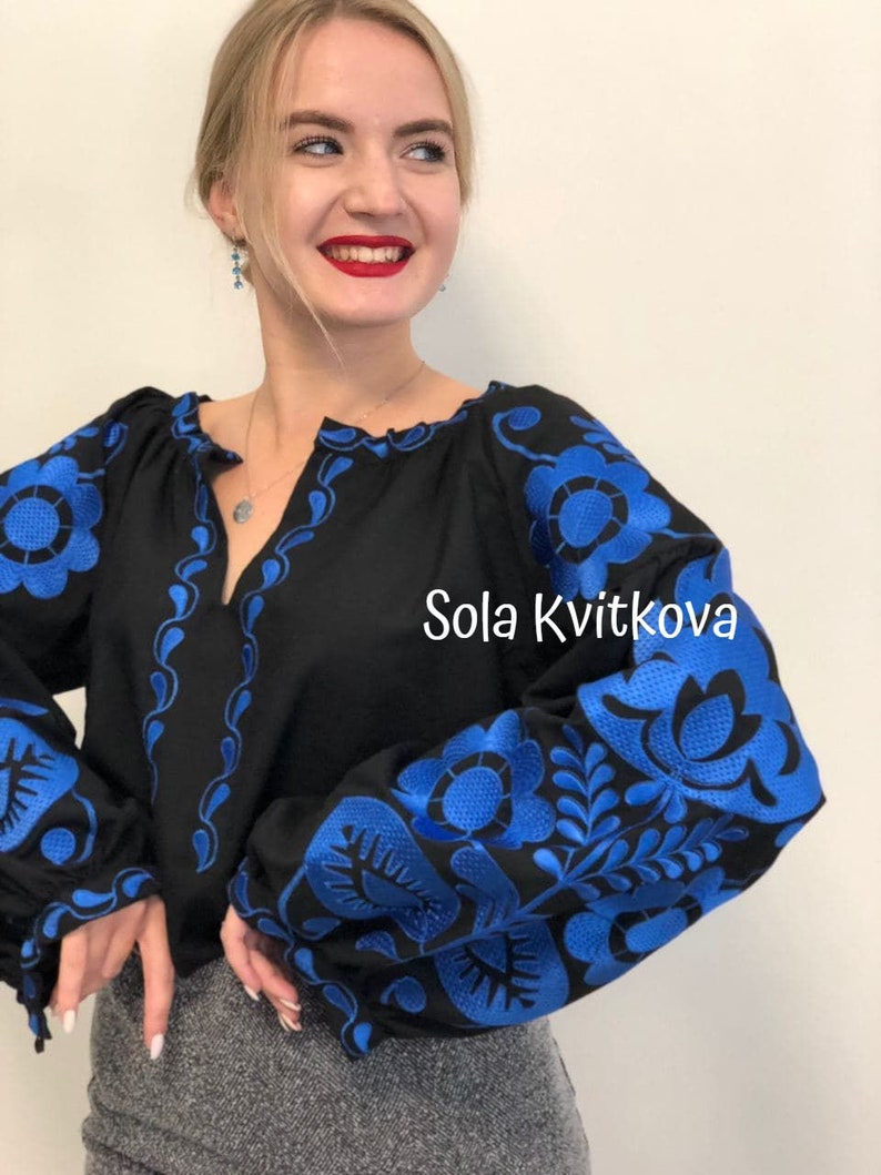 Op de afbeelding: Een zwarte geborduurde blouse met blauwe bloemenmotieven. De tekst "Sola Kvitkova" is zichtbaar aan de rechterkant van de blouse.