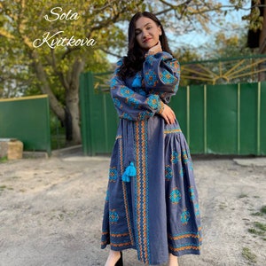 May include: A woman wearing a long, blue embroidered dress with orange and blue geometric patterns. The dress has a high neckline and long sleeves. The woman is standing in front of a green gate and a tree.