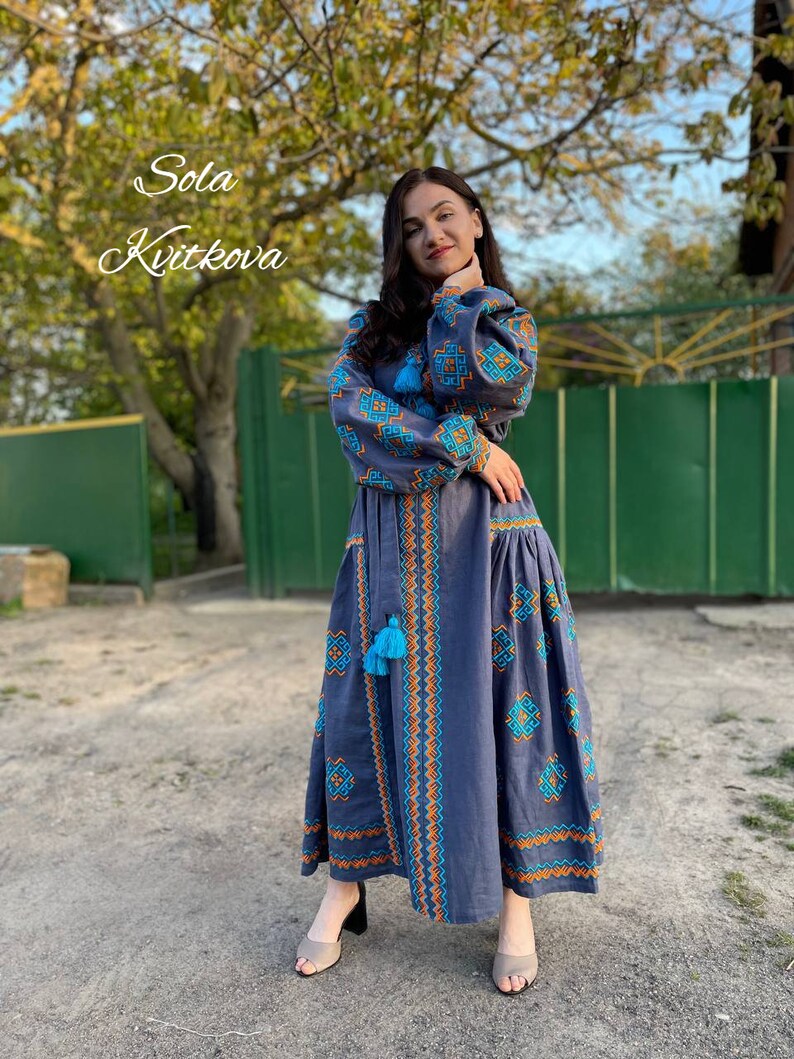 May include: A woman wearing a long, blue embroidered dress with orange and blue geometric patterns. The dress has a high neckline and long sleeves. The woman is standing in front of a green gate and a tree.