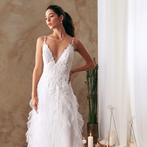 K&ouml;nnte beinhalten: Ein wei&szlig;es Brautkleid mit einem Spitzenoberteil und einem T&uuml;llrock mit R&uuml;schen. Das Kleid hat einen V-Ausschnitt und Spaghettitr&auml;ger.