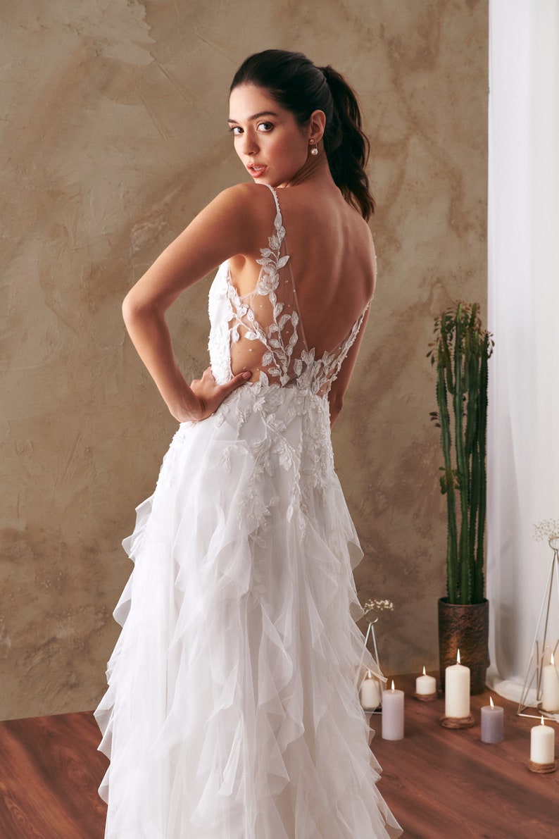 K&ouml;nnte beinhalten: Ein wei&szlig;es Brautkleid mit einem Spitzenoberteil und einem flie&szlig;enden T&uuml;llrock. Das Kleid hat einen tiefen R&uuml;cken und zarte florale Stickereien.