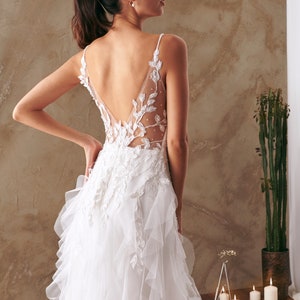 K&ouml;nnte beinhalten: Ein wei&szlig;es Brautkleid mit tiefem R&uuml;cken und einem R&uuml;schenrock. Das Kleid ist mit zarten Blumenstickereien und einem durchsichtigen Einsatz verziert.