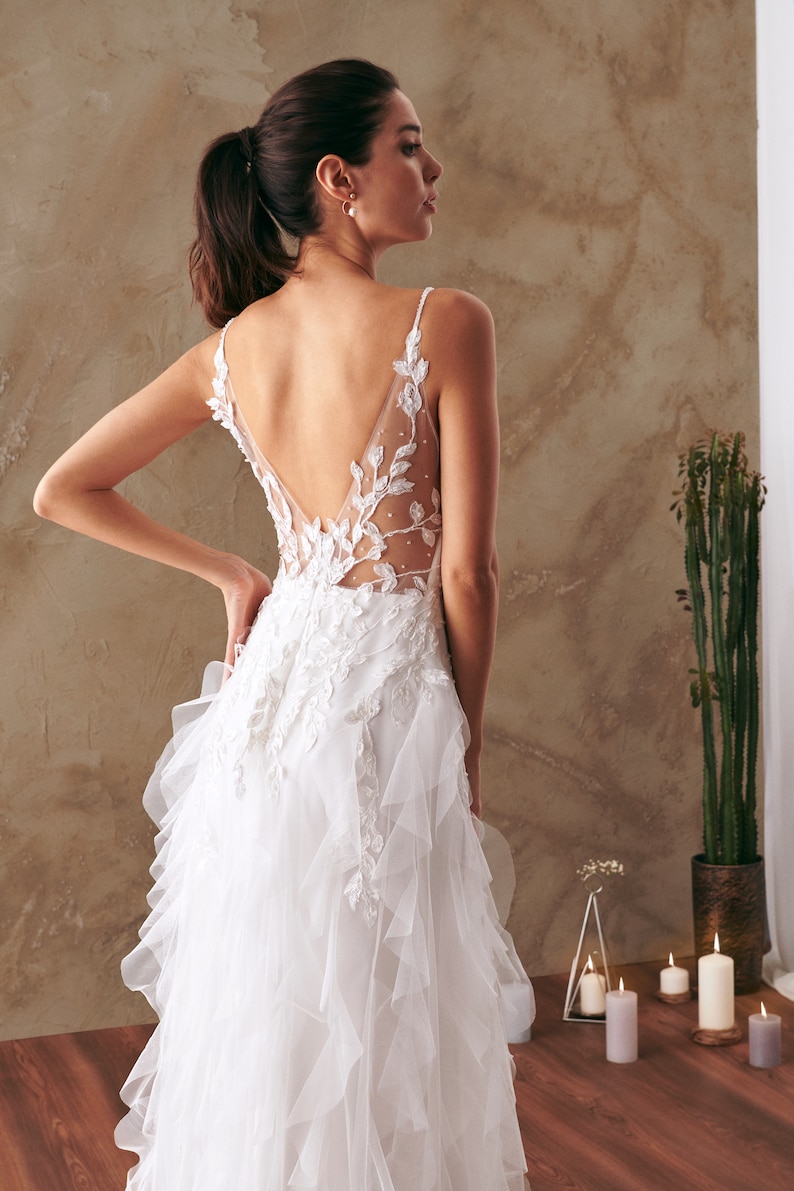 K&ouml;nnte beinhalten: Ein wei&szlig;es Brautkleid mit tiefem R&uuml;cken und einem R&uuml;schenrock. Das Kleid ist mit zarten Blumenstickereien und einem durchsichtigen Einsatz verziert.