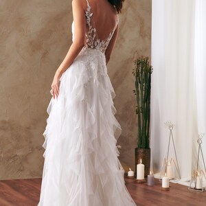 K&ouml;nnte beinhalten: Ein wei&szlig;es Brautkleid mit einem Spitzenoberteil und einem T&uuml;llrock mit mehreren R&uuml;schenlagen. Das Kleid hat einen tiefen R&uuml;cken mit einem Blumendesign.