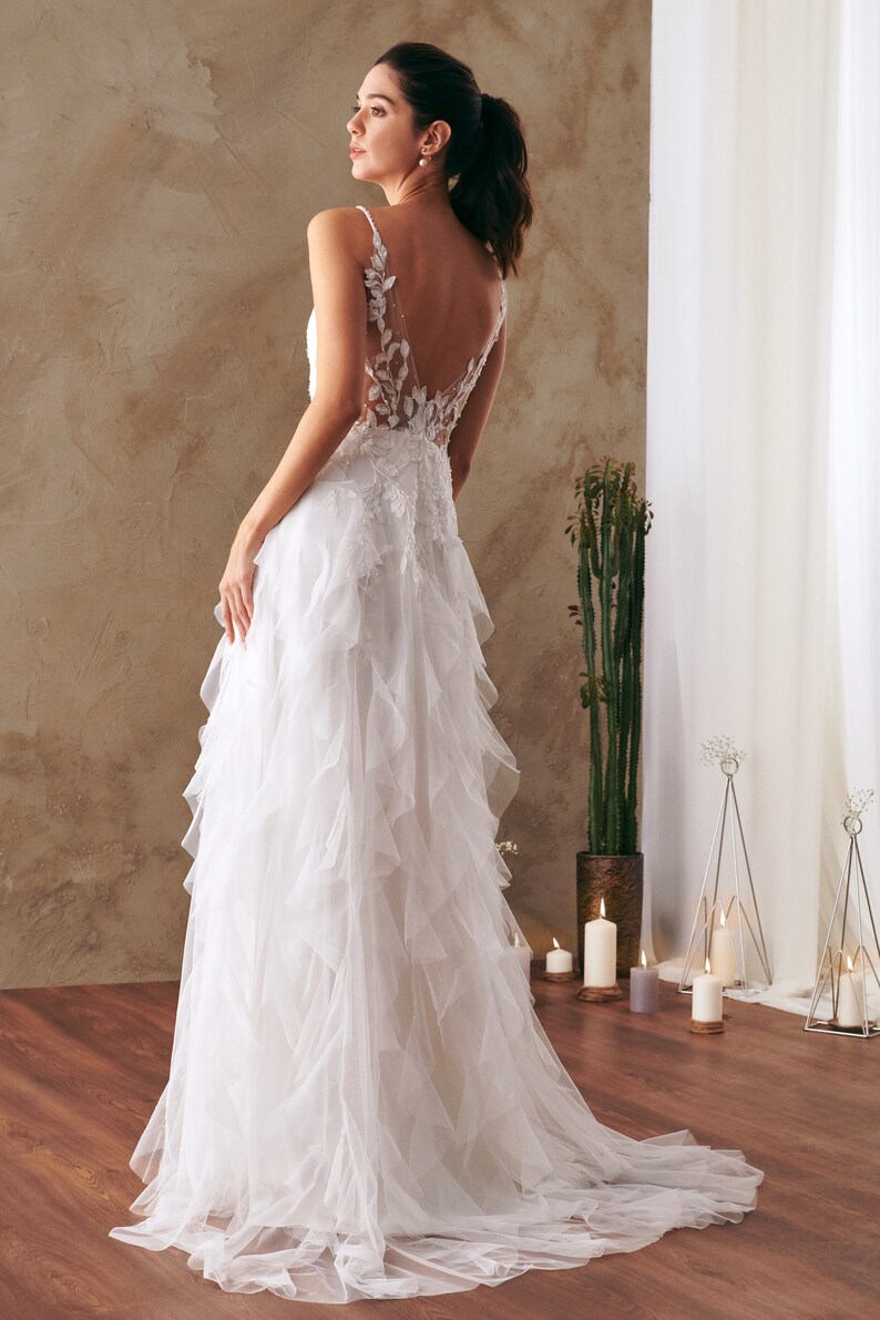 K&ouml;nnte beinhalten: Ein wei&szlig;es Brautkleid mit einem Spitzenoberteil und einem T&uuml;llrock mit mehreren R&uuml;schenlagen. Das Kleid hat einen tiefen R&uuml;cken mit einem Blumendesign.