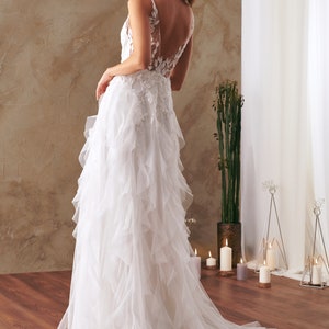 K&ouml;nnte beinhalten: Ein wei&szlig;es Brautkleid mit einem Spitzenoberteil und einem flie&szlig;enden T&uuml;llrock mit mehreren R&uuml;schenlagen. Das Kleid hat einen tiefen R&uuml;cken und ein schlichtes, elegantes Design.