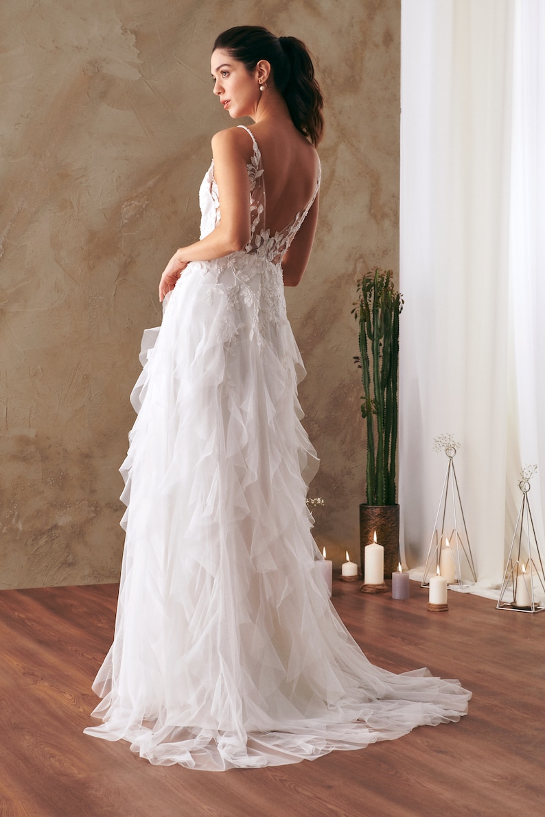 K&ouml;nnte beinhalten: Ein wei&szlig;es Brautkleid mit einem Spitzenoberteil und einem flie&szlig;enden T&uuml;llrock mit mehreren R&uuml;schenlagen. Das Kleid hat einen tiefen R&uuml;cken und ein schlichtes, elegantes Design.