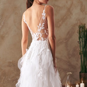 K&ouml;nnte beinhalten: Ein wei&szlig;es Brautkleid mit einem Spitzenoberteil und einem T&uuml;llrock mit R&uuml;schen. Das Kleid hat einen tiefen R&uuml;cken mit einem Blumendesign.