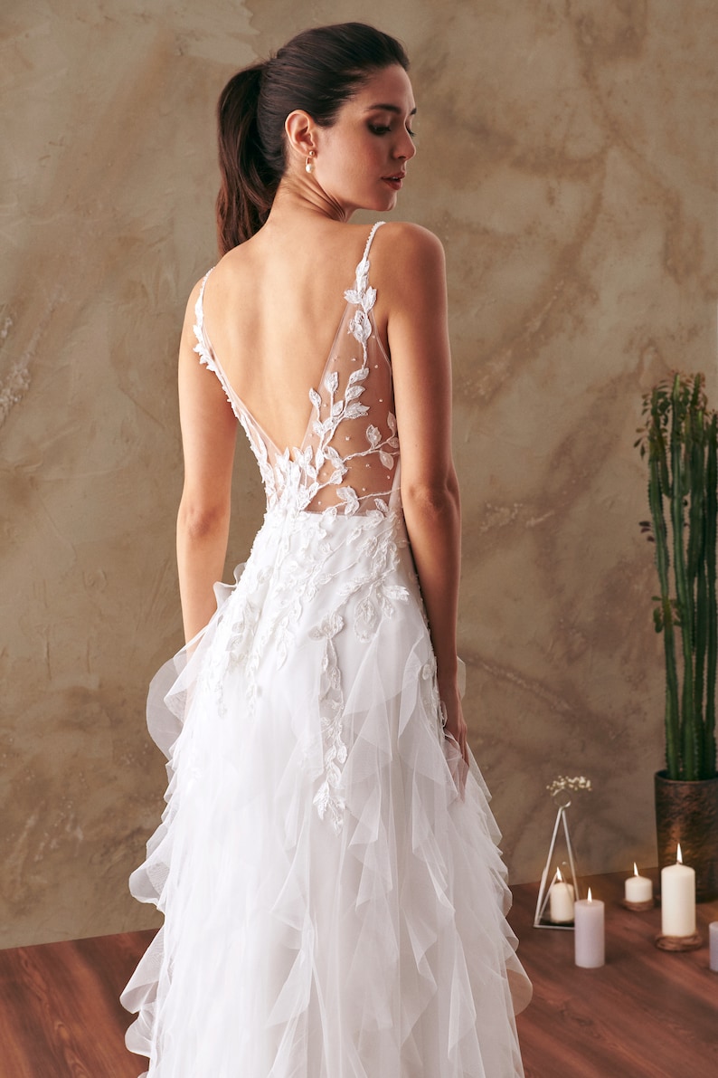 K&ouml;nnte beinhalten: Ein wei&szlig;es Brautkleid mit einem Spitzenoberteil und einem T&uuml;llrock mit R&uuml;schen. Das Kleid hat einen tiefen R&uuml;cken mit einem Blumendesign.