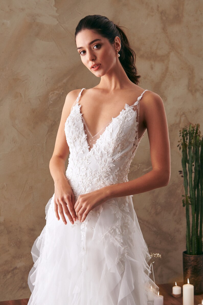 K&ouml;nnte beinhalten: Ein wei&szlig;es Brautkleid mit einem Spitzenoberteil und einem R&uuml;schenrock. Das Kleid hat einen V-Ausschnitt und d&uuml;nne Tr&auml;ger.