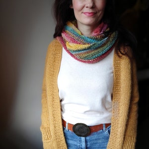 May include: A woman wearing a mustard yellow cardigan with a multi-coloured scarf, a white t-shirt, blue jeans, and a brown belt with a circular buckle.