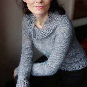 Może przedstawiać: Jaszek z dzianiny w jasnoniebieskim kolorze z delikatnym wzorem koronkowym wokół dekoltu. Sweter wykonany jest z miękkiej, lekkiej przędzy i ma luźny krój.