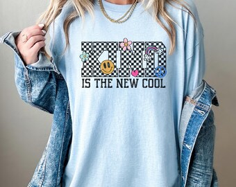 May include: Light blue long-sleeve shirt with the word "KIND" in a checkered pattern, featuring a smiley face, flowers, a rainbow, and a peace sign. The text "IS THE NEW COOL" is below the word "KIND".