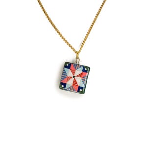 May include: A gold-colored chain necklace with a square pendant. The pendant features a colorful geometric design with red, blue, and white shapes. The pendant is framed with a green border, creating a striking contrast against the white background.