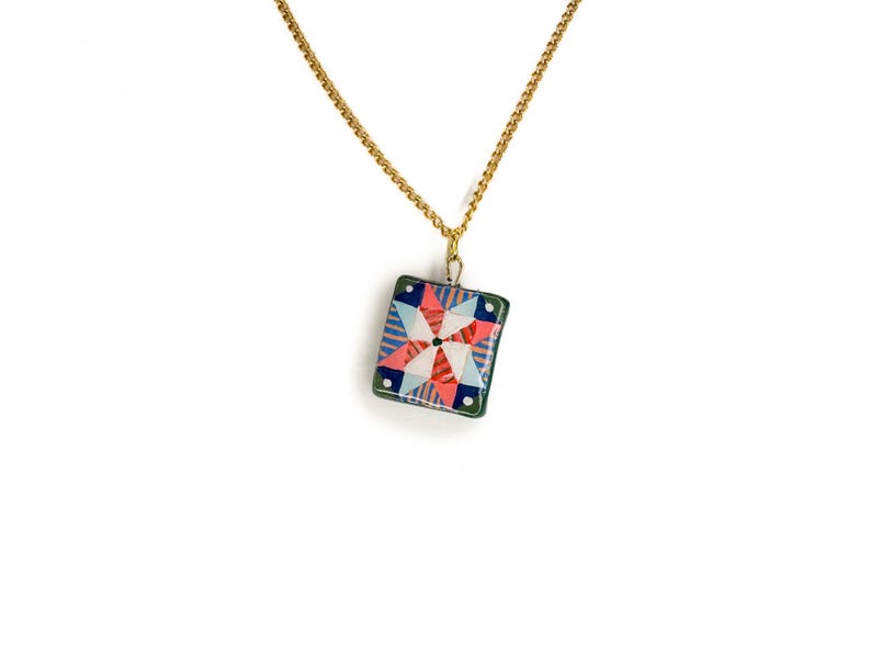 May include: A gold-colored chain necklace with a square pendant. The pendant features a colorful geometric design with red, blue, and white shapes. The pendant is framed with a green border, creating a striking contrast against the white background.