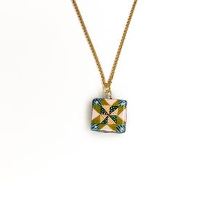 May include: A square pendant necklace with a gold chain. The pendant features a colorful patchwork design with pink, green, blue, and yellow patterns. The necklace is displayed against a white background.