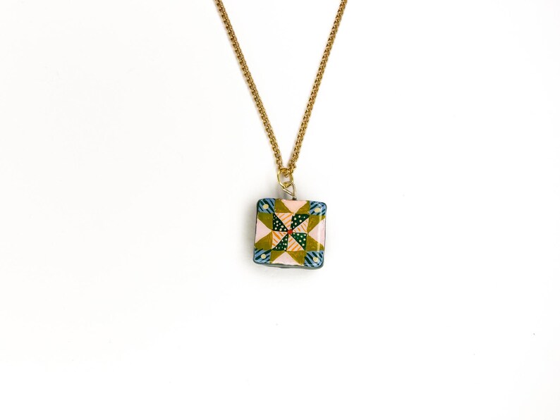 May include: A square pendant necklace with a gold chain. The pendant features a colorful patchwork design with pink, green, blue, and yellow patterns. The necklace is displayed against a white background.