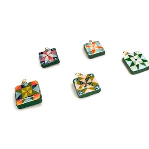 May include: Five square pendants with colorful quilt patterns and gold-colored loops. The pendants have a dark green border and feature various geometric designs in shades of red, orange, green, and blue. They are set against a white background.