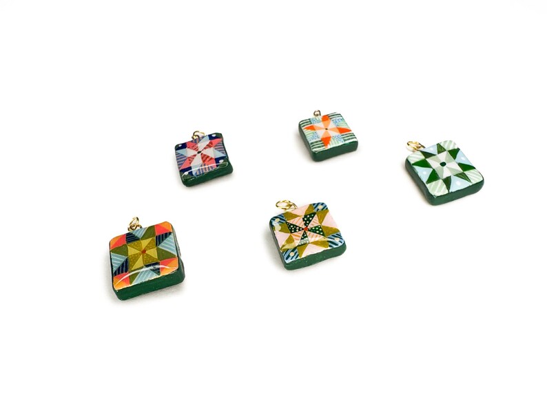 May include: Five square pendants with colorful quilt patterns and gold-colored loops. The pendants have a dark green border and feature various geometric designs in shades of red, orange, green, and blue. They are set against a white background.