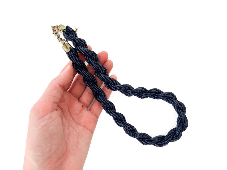 May include: A dark blue beaded necklace with a braided design. The necklace is made of small, round beads that are woven together to create a rope-like effect.