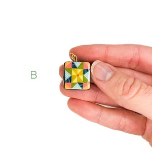May include: A square pendant with a colorful geometric design, held in a hand. The pendant features a patchwork quilt pattern in shades of yellow, green, pink, and blue, with a gold-colored bail.