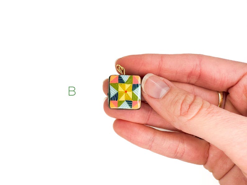 May include: A square pendant with a colorful geometric design, held in a hand. The pendant features a patchwork quilt pattern in shades of yellow, green, pink, and blue, with a gold-colored bail.