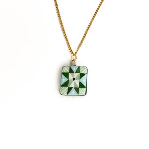 May include: A gold-colored chain necklace with a rectangular pendant. The pendant features a geometric design in shades of green, light blue, and white. The design is centered with a dark green square. The necklace is displayed against a white background.