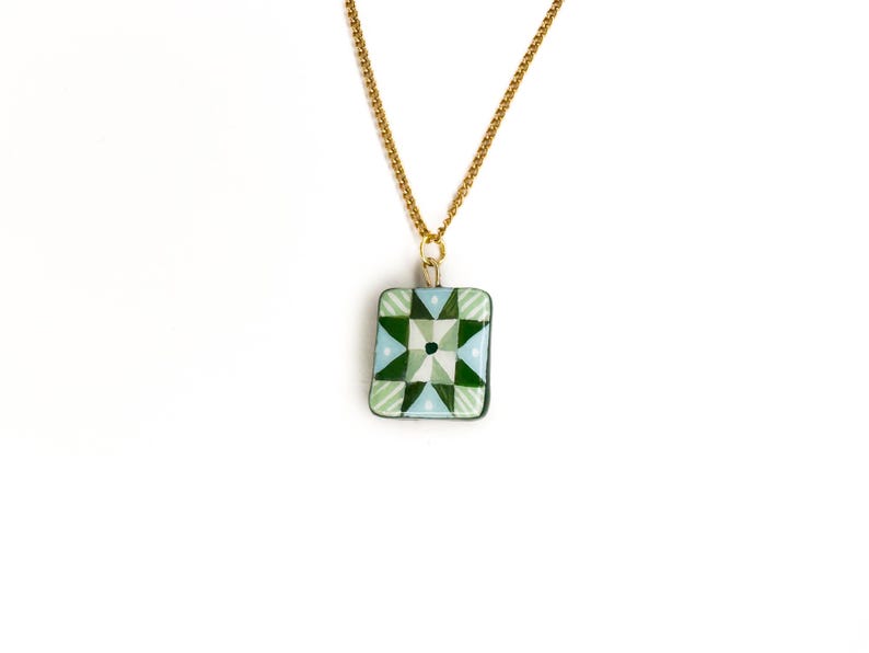 May include: A gold-colored chain necklace with a rectangular pendant. The pendant features a geometric design in shades of green, light blue, and white. The design is centered with a dark green square. The necklace is displayed against a white background.