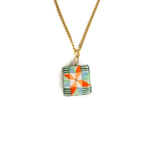May include: A gold-colored chain necklace with a square pendant. The pendant features a colorful patchwork design with orange, green, and blue geometric shapes. The necklace is displayed against a white background.